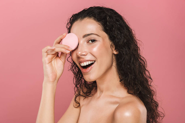 Beauty portrait of a cheerful young topless woman with curly brunette hair over pink background, wiping face with a cosmetic sponge - Fotografie, Obrázek
