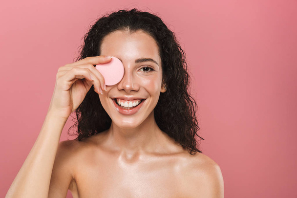 Schönheitsporträt einer lebenslustigen jungen Frau oben ohne mit lockigem brünetten Haar vor rosa Hintergrund, die das Gesicht mit einem Kosmetikschwamm wischt - Foto, Bild