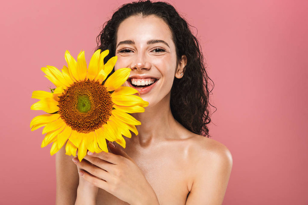 Photo of beautiful nude woman 20s with long hair smiling and holding yellow sunflower isolated over pink background - Photo, image