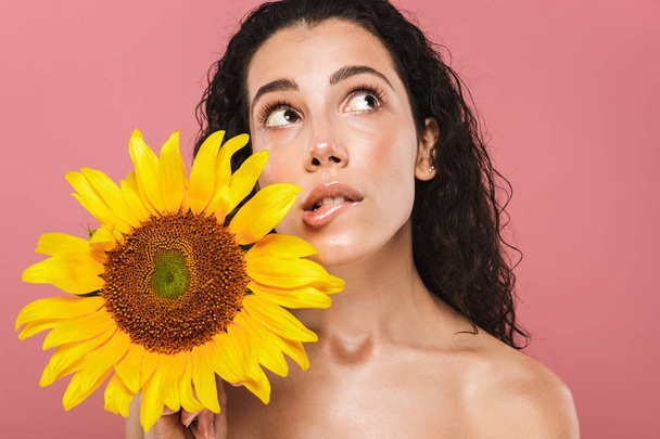 Beauty portrait of a lovely young topless woman with curly brunette hair posing over pink background, holding a sunflower - Foto, afbeelding