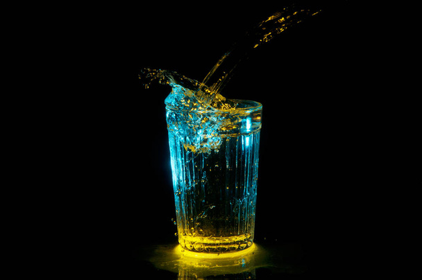 Pouring liquid into an old glass and making a wave under yellow and blue lights isolated on a black background - Photo, Image