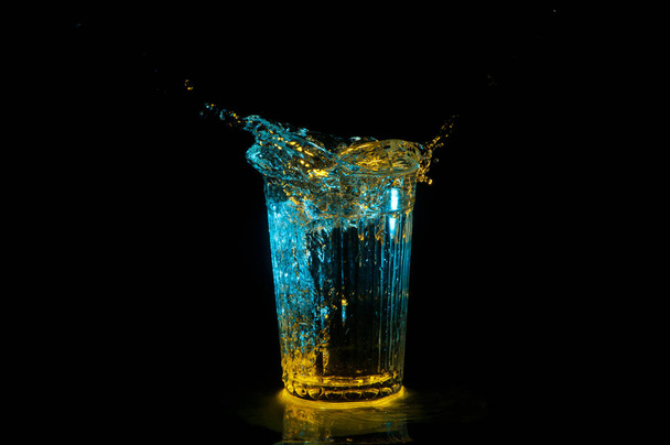 Ellipsi vettä roiskuu ulos harjattu lasi keltaisen ja sinisen valon eristetty mustalla pohjalla
 - Valokuva, kuva