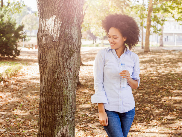 Portrait d'une jeune femme noire souriante tenant une bouteille d'eau dans la nature, coiffure afro, chemise bleue, éclat de soleil en arrière-plan
 - Photo, image