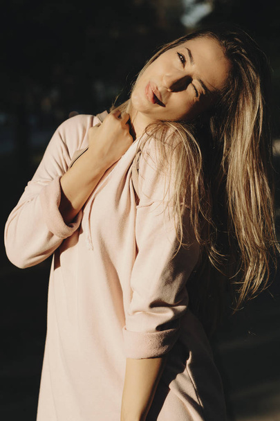 Coquettish young blond woman in pink sweatshirt standing in sunshine having fun and showing tongue  - Фото, изображение