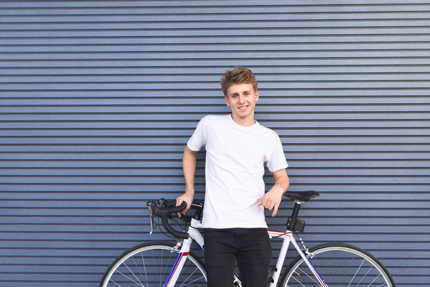 Retrato de un tipo feliz con una camiseta blanca de pie con una bicicleta blanca contra la pared, mirando a la cámara y sonriendo. Estudiante feliz con un retrato de bicicleta
. - Foto, Imagen