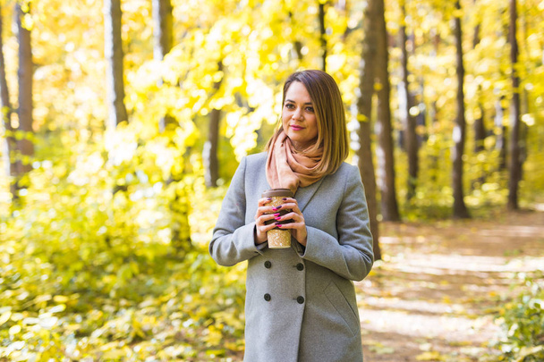 Φθινόπωρο, φύση και άνθρωποι έννοια - νεαρή όμορφη γυναίκα σε γκρι παλτό, κρατώντας ένα φλιτζάνι καφέ - Φωτογραφία, εικόνα
