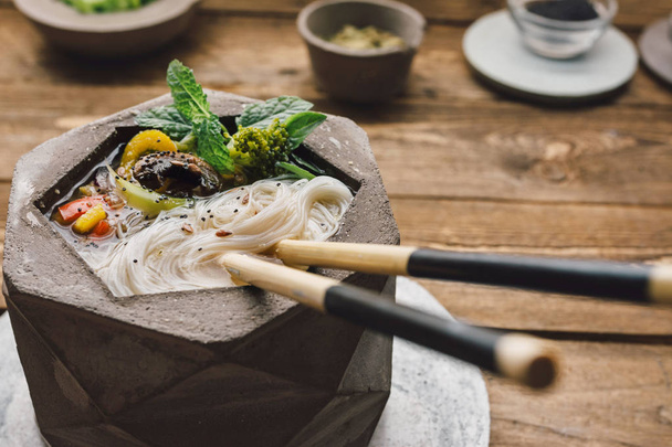Tigela geométrica de sopa de macarrão asiático com pauzinhos, em mesa de madeira e legumes frescos
 - Foto, Imagem