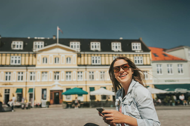 Bella giovane donna seduta per strada nel centro della città in una giornata di sole e con il cellulare in mano e sorridente
. - Foto, immagini