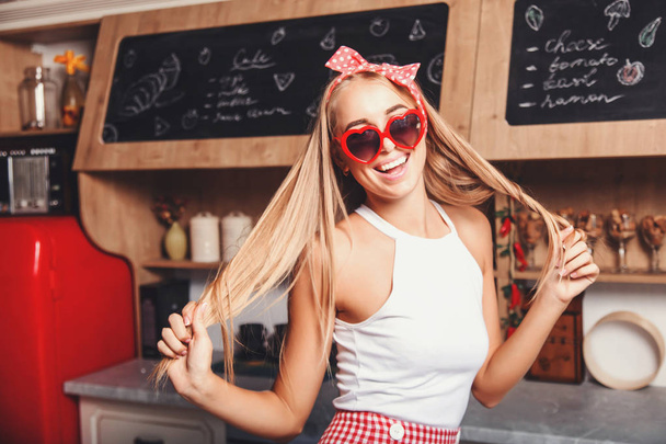 Pretty blonde long hair housewife wearing hearts-sunglasses dancing and having fun in the red loft comfortable sunny kitchen - 写真・画像