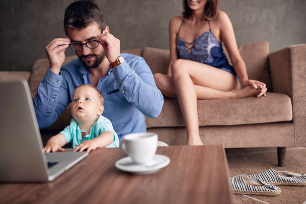 Busy father working with computer while looking after his baby son - 写真・画像