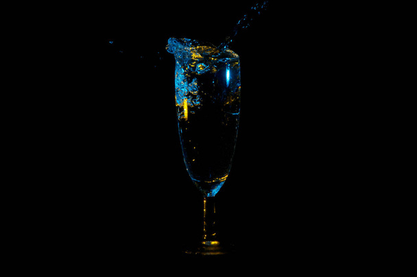 blaue Flüssigkeit mit gelben Reflexen, die aus einem Weinglas auf schwarzem Hintergrund herausspritzen - Foto, Bild