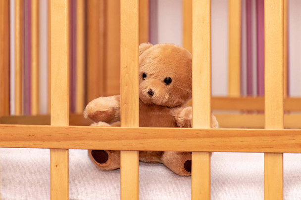 Toy teddy bear in a baby cot. A teddy bear sits behind a wooden strap. Children room - Foto, afbeelding