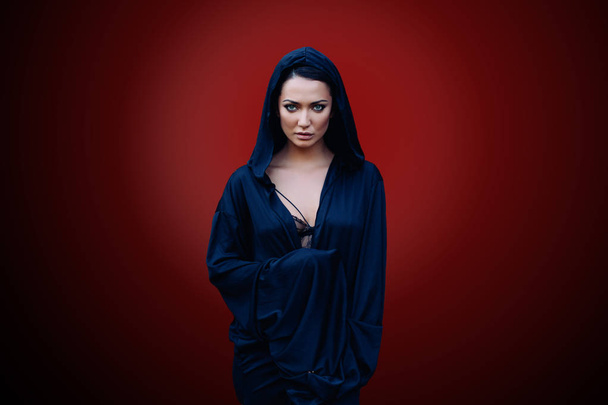 Young beautiful woman with a black hair and in the dark blue cloak with hood at the red background - Fotó, kép