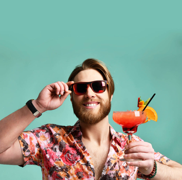 Jeune homme au chapeau montrer pouce vers le haut boire du jus de boisson cocktail margarita rouge heureux en regardant la caméra sur bleu
 - Photo, image