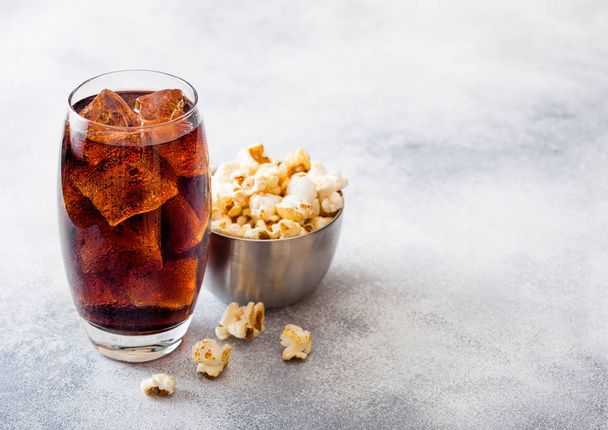 Glass of cola soda drink with ice cubes and steel bowl of popcorn snack on stone kitchen background. - Valokuva, kuva