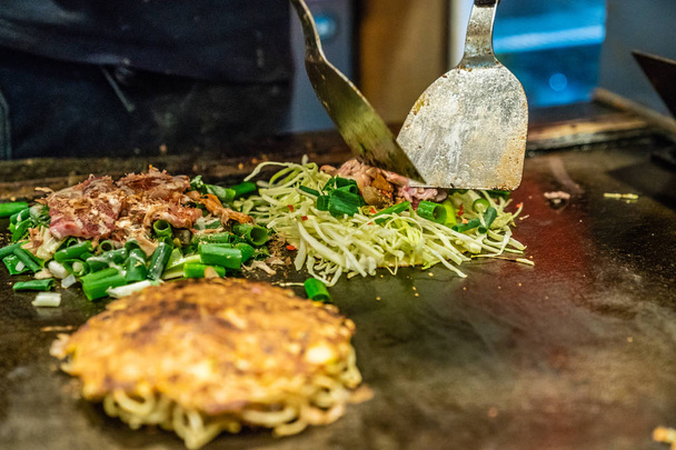 chef is using the spatula to cook okonomiyaki on the iron plate, he puts some slices meat and vegetables in the food - Fotoğraf, Görsel