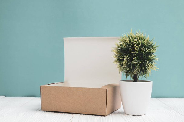 Empty Package brown cardboard box or tray and little decoration tree in white vase on bright white wooden table with green wall background. copy space for add text or advertising word. - Foto, Bild