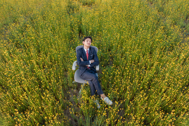 Aasialainen liikemies makaa tuolilla suurella keltaisella kukkakentällä
 - Valokuva, kuva