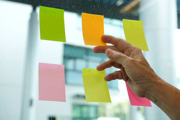 ręka trzymać notatki kleju na ścianie szkło w miejscu pracy. Karteczkę papieru harmonogramu przypomnienia dla kreatywnych idei & biznes burza mózgów - Zdjęcie, obraz
