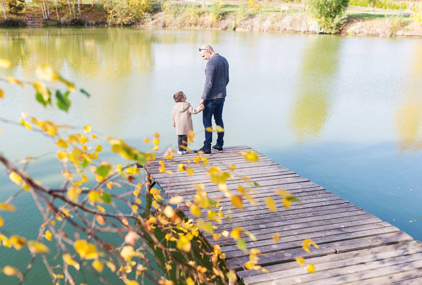 Young bald father and her little boy stand on the wooden pier in warm autumn day. Back view - Photo, Image