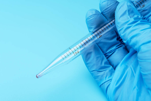 Een gehandschoende hand met een laboratorium pipet op een blauwe achtergrond. - Foto, afbeelding