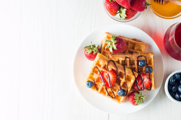 Photo of fresh homemade food made of berry Belgian waffles with honey, chocolate, strawberry, blueberry, maple syrup and cream. Healthy dessert breakfast concept with juice.  - Photo, Image