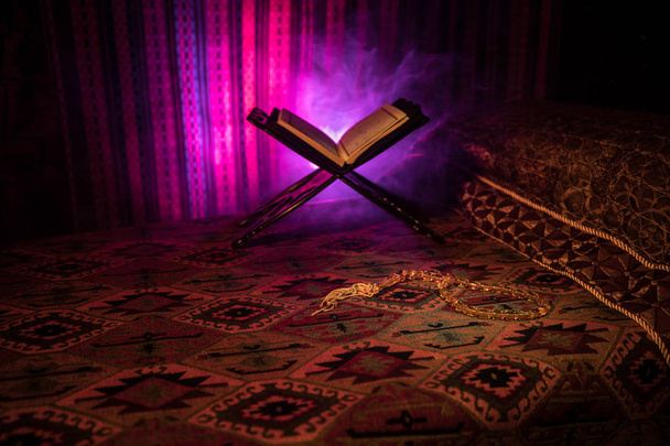 Ανοίξτε το ιερό βιβλίο των μουσουλμάνων σε στάση στο ανατολικό χαλί με σκούρο τονισμένο θολό φόντο. Μουσουλμανική θρησκεία. Επιλεκτική εστίαση. Εσωτερική λήψη. - Φωτογραφία, εικόνα