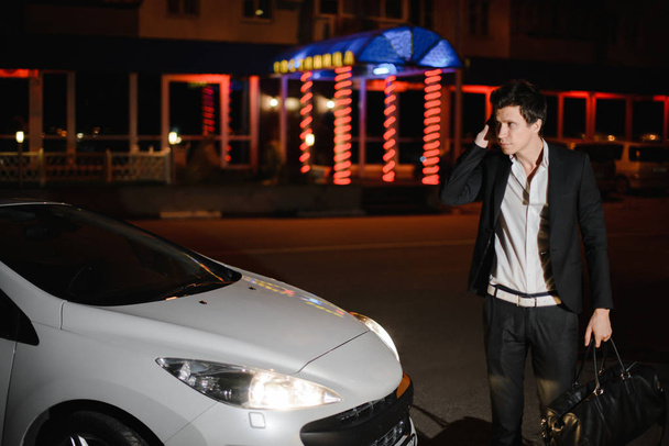 Portrait of a handsome man standing next to his white cabriolet. Nightlife. Businessman in suit in luxury car. - Fotó, kép