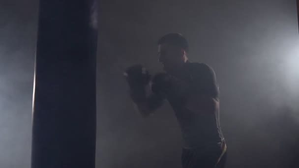 Boxer hits punching bag in dark gym in slow motion. Young man training indoors - Filmagem, Vídeo