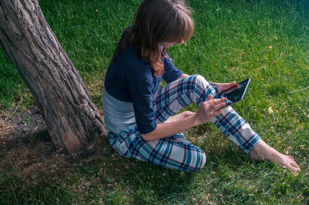 Jonge roodharige vrouw in chekered slijtage met een tablet-pc in het park leunend tegen een oude boom, zoals ze wordt geschoven met behulp van het touchscreen - Foto, afbeelding
