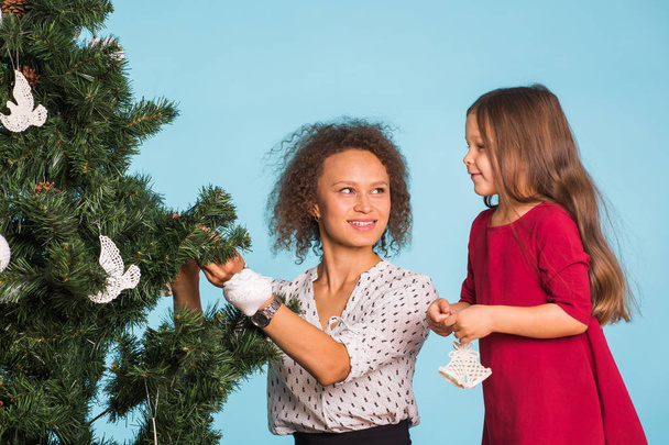 Urlaub, Familie und Weihnachtskonzept - hübsches kleines Mädchen mit gemischter Rasse Mutter schmücken einen Weihnachtsbaum auf blauem Hintergrund - Foto, Bild