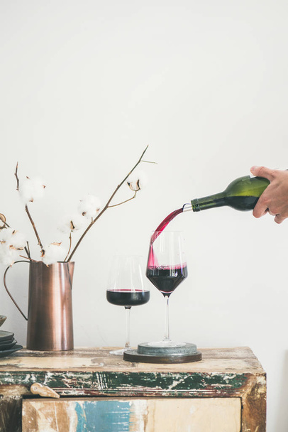 Red wine pouring from bottle into wineglass over rustic kitchen countertop, white background behind - Φωτογραφία, εικόνα