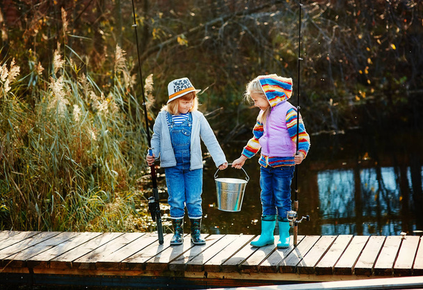 Two little girls with fishing rods, they stand on a wooden pontoon and look at the fish caught in a bucket - Фото, зображення