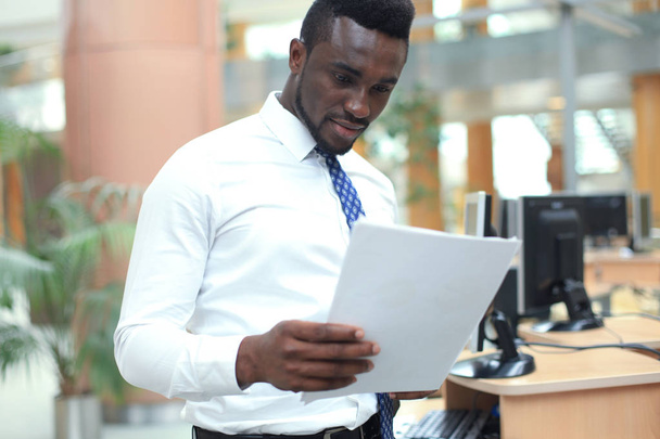 Successo afroamericano uomo d'affari lettura carta sul posto di lavoro, guardando documento finanziario, in possesso di caffè
. - Foto, immagini