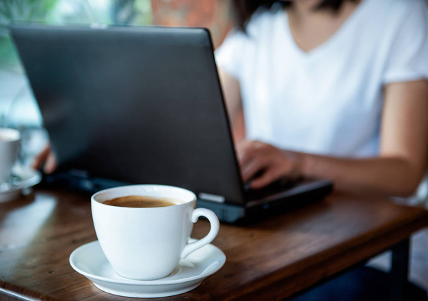 Kaffeetasse auf Holztisch mit verschwommenem Mädchen mit Laptop. - Foto, Bild