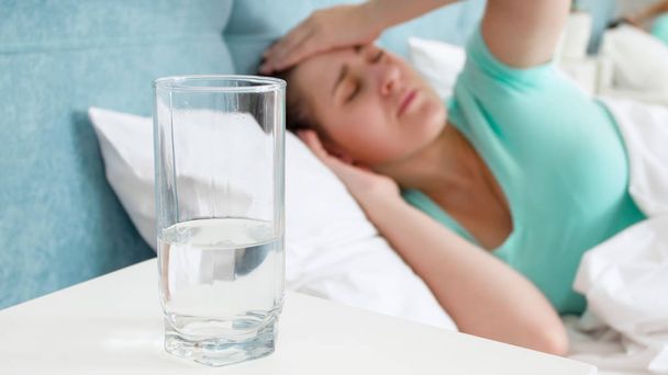 Immagine di una donna malata che soffre di mal di testa sdraiata a letto e di un bicchiere d'acqua sul comodino
 - Foto, immagini