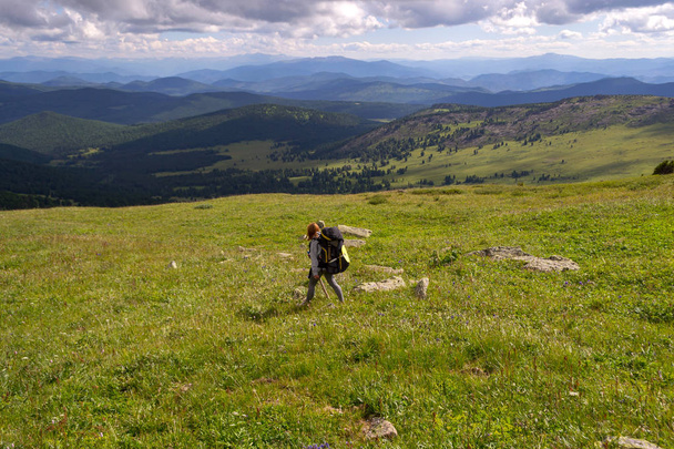 Una chica turista en una capucha, con una gran mochila y un palo de madera en sus manos desciende de la montaña a las rocas con musgo en frente de ella es un valle verde con colinas cielo azul con nubes blancas
 - Foto, imagen