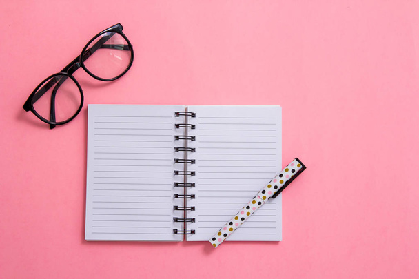 Notepad, pen and glasses on pink background. Flat ley composition. Office supplies on the table. - Фото, зображення