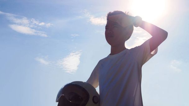 Asijský mladík drží helmu a zdravit se silným slunečním záře v pozadí. (vybrané zaměření) - Fotografie, Obrázek