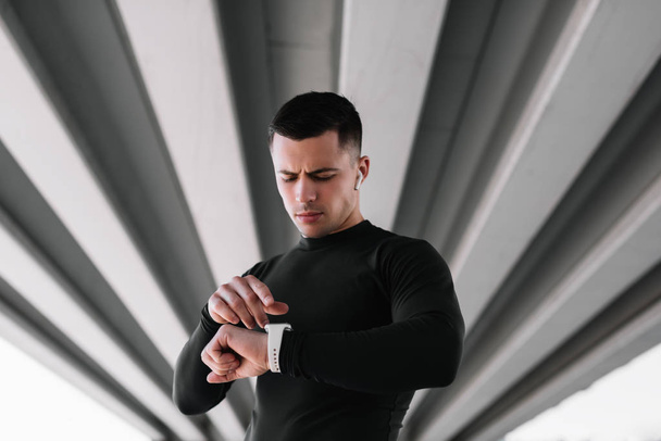 Portrait of muscular athlete listening music wearing wireless headphones and using smartwatch standing on the street . Professional sportsman or runner  in fitted suit ready to workout. Training. - Photo, Image