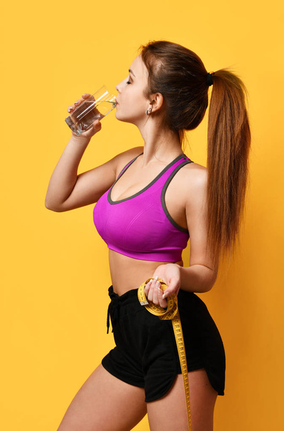 Beautiful sport woman hold tape measure and drinking water from glass on yellow background - Фото, зображення