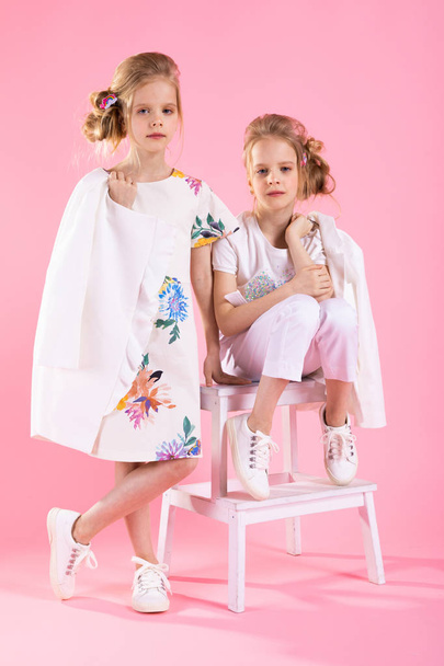 Twins dziewczyny w jasne ubrania pozowanie w pobliżu schodów z dwa kroki na różowym tle. - Zdjęcie, obraz
