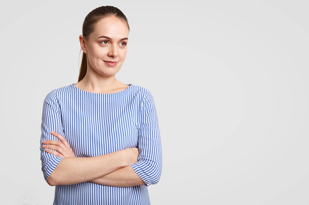 Horizontal shot of thoughtful European female with pony tail, keeps hands crossed, dressed in striped blouse, poses against white background with copy space aside for your promotion or advertisement - Фото, зображення