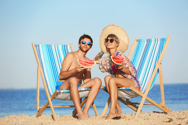 Young couple with watermelon slices in beach chairs at seacoast - Zdjęcie, obraz