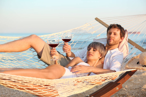 Young couple resting with glasses of wine in hammock on beach - 写真・画像