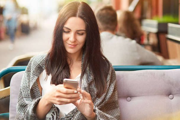 Online internet connection concept. Beautiful young brunette lady uses mobile phone for searching internet, wrapped in coverlet, spends free time in coffee shop, recieves message from boyfriend - 写真・画像