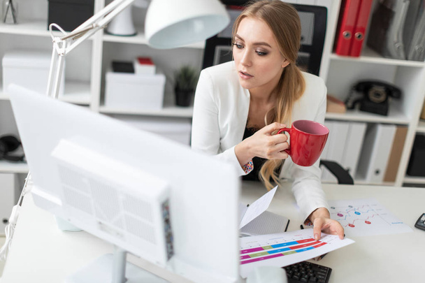 Mladá dívka sedí u počítače, recepce v kanceláři a drží pohár a dokumenty. Před dívku na stůl jsou diagramy. - Fotografie, Obrázek