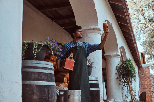 Birraio esperto in grembiule in piedi all'aperto controllando la qualità della bevanda prodotta presso la fabbrica di birra
. - Foto, immagini
