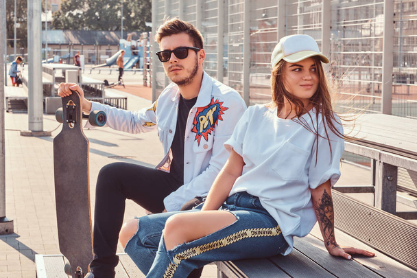 Atractive pár divatos öltözött fiatal hippik ül a padon a városi sportkomplexum, napsütéses napon gördeszka. - Fotó, kép