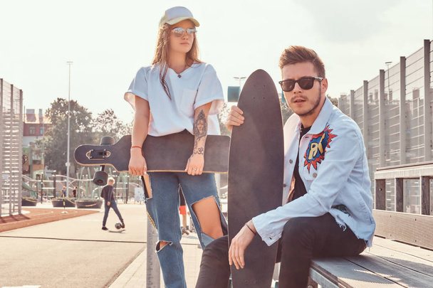 Close-up portrait of young trendy dressed hipster couple with skateboards posing at city sports complex on sunny day - Photo, Image